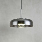 Smoke grey glass Clérachic pendant light against a grey plaster wall, its silver-toned stem and lit disc visible inside.