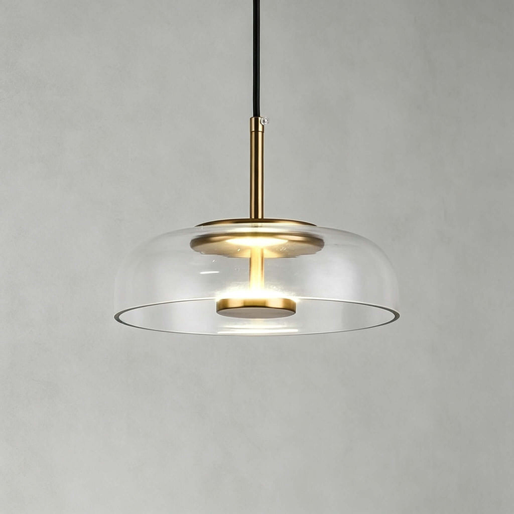 Clear glass Clérachic pendant light on a soft grey wall, with a brass stem and a warm LED disc glowing within.