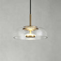 Clear glass Clérachic pendant light on a soft grey wall, with a brass stem and a warm LED disc glowing within.