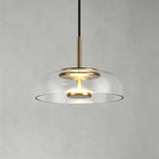 Clear glass Clérachic pendant light on a soft grey wall, with a brass stem and a warm LED disc glowing within.
