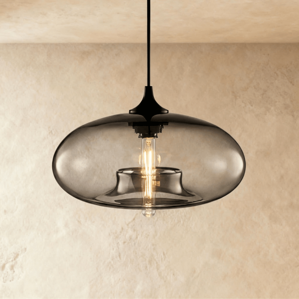 Clérachic pendant light with a smoked-grey blown-glass oval shade over a black cord and cap, filament bulb lit, on a limewash wall.
