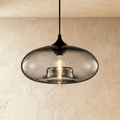 Clérachic pendant light with a smoked-grey blown-glass oval shade over a black cord and cap, filament bulb lit, on a limewash wall.
