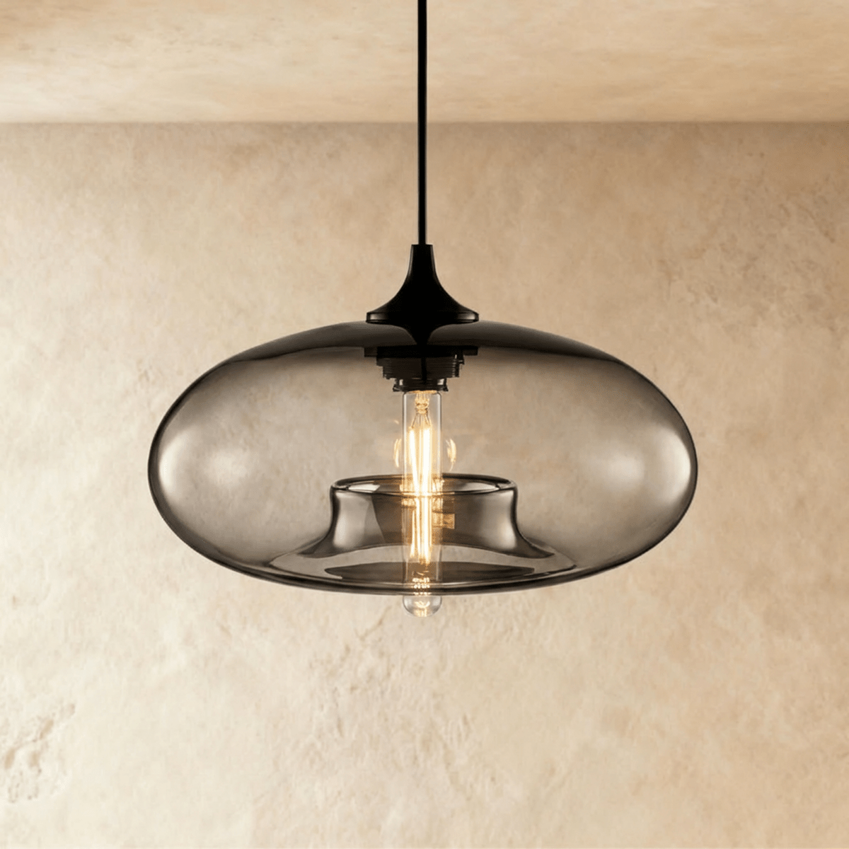 Clérachic pendant light with a smoked-grey blown-glass oval shade over a black cord and cap, filament bulb lit, on a limewash wall.