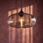 Clérachic pendant light close-up, a clear-to-smoke oval glass shade with a glowing tube bulb against a pink plaster wall.