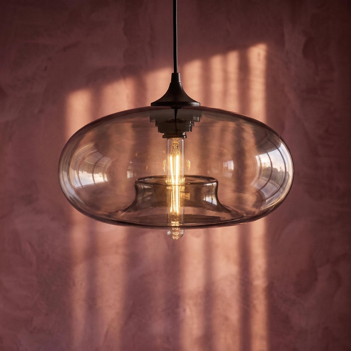 Clérachic pendant light close-up, a clear-to-smoke oval glass shade with a glowing tube bulb against a pink plaster wall.