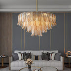 A wide Clérachic leaf-glass pendant in amber hangs over a pale sofa, against a grey wall with slim gold lines.