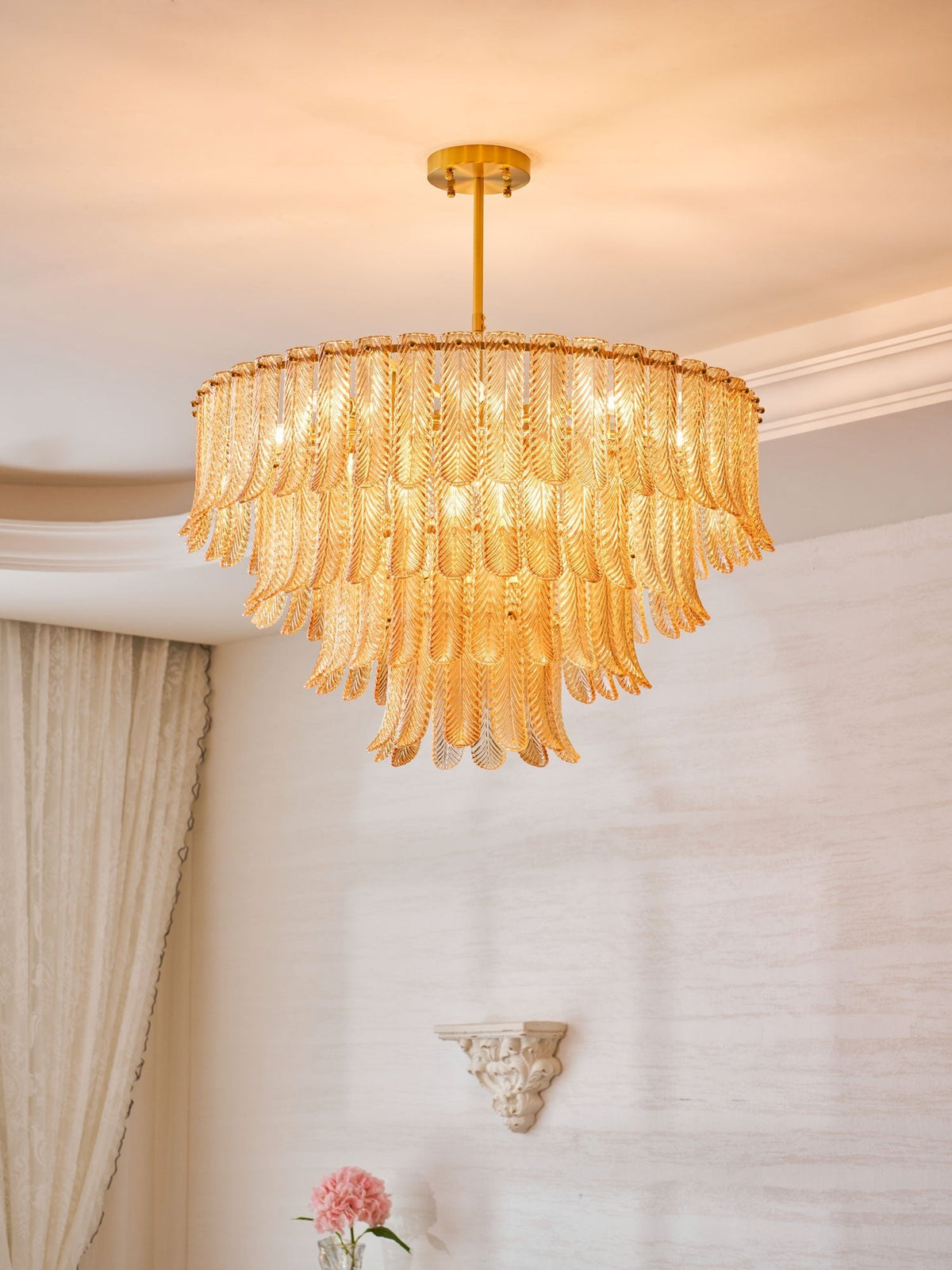 Looking up at a Clérachic tiered pendant of amber glass leaves, lit warm, hanging from a brass rod beside a plaster ceiling edge.
