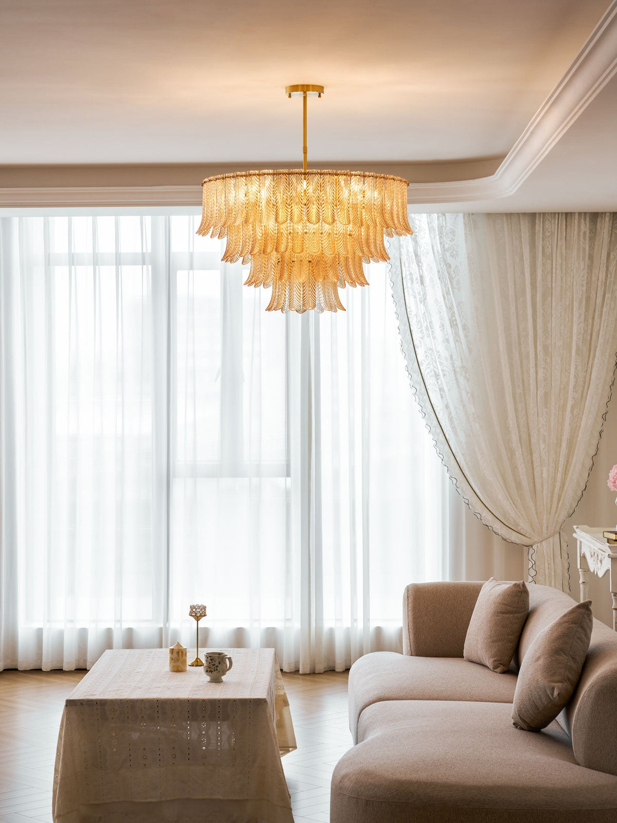 A Clérachic pendant of layered glass leaves hangs high above a linen-covered table and a taupe sofa, in front of sheer curtains.