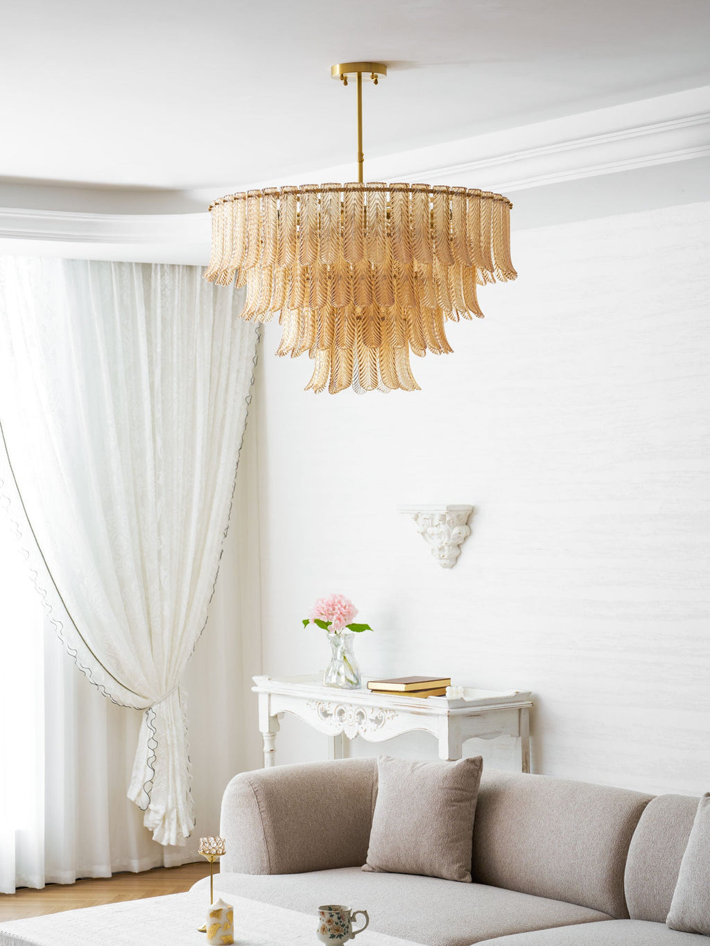 A Clérachic amber leaf-glass pendant hangs in a white living room, with a sofa, a carved console and pink flowers beyond.