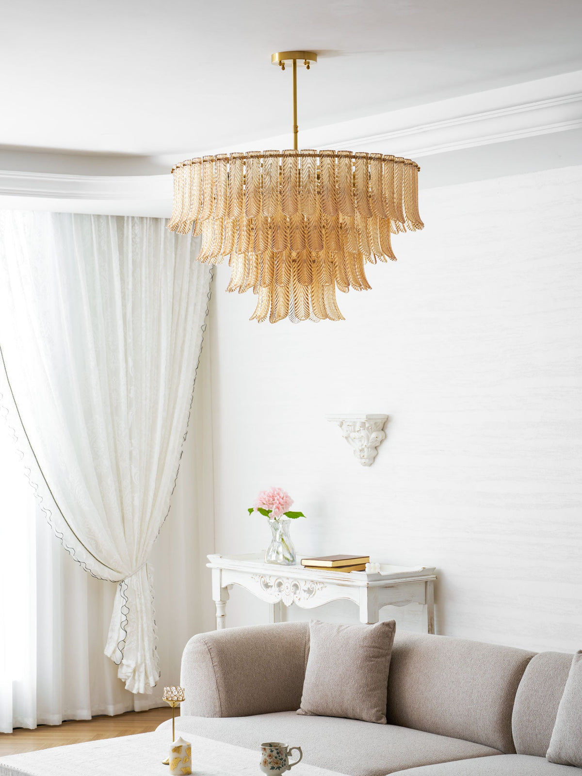 A Clérachic amber leaf-glass pendant hangs in a white living room, with a sofa, a carved console and pink flowers beyond.