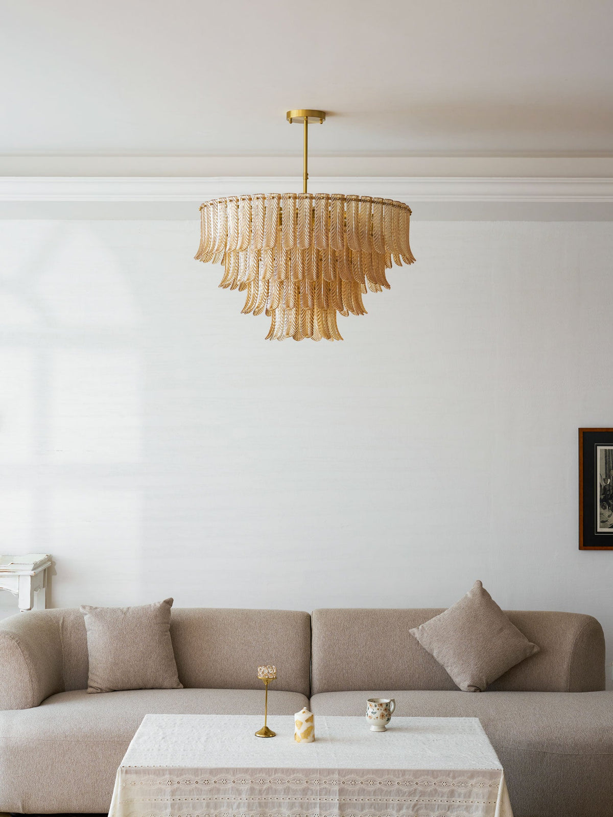 Seen from across a white room, a Clérachic leaf-glass pendant hangs above a taupe sofa and a lace-covered table.