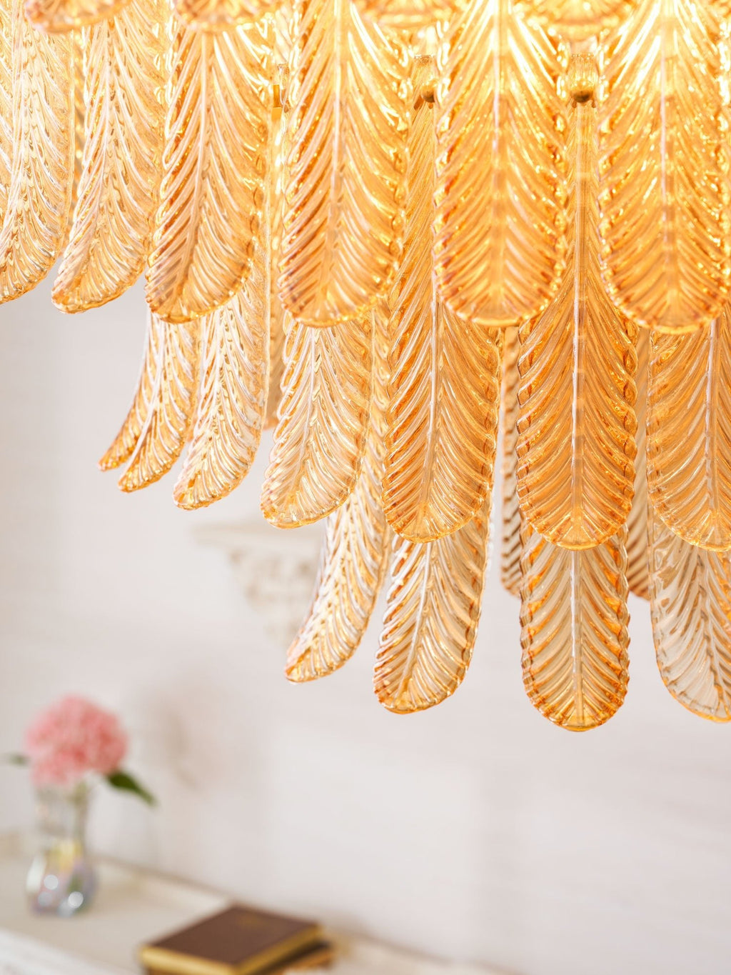 Close view of the overlapping amber glass leaves on this Clérachic pendant, with a vase of pink flowers blurred below.
