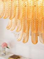 Close view of the overlapping amber glass leaves on this Clérachic pendant, with a vase of pink flowers blurred below.