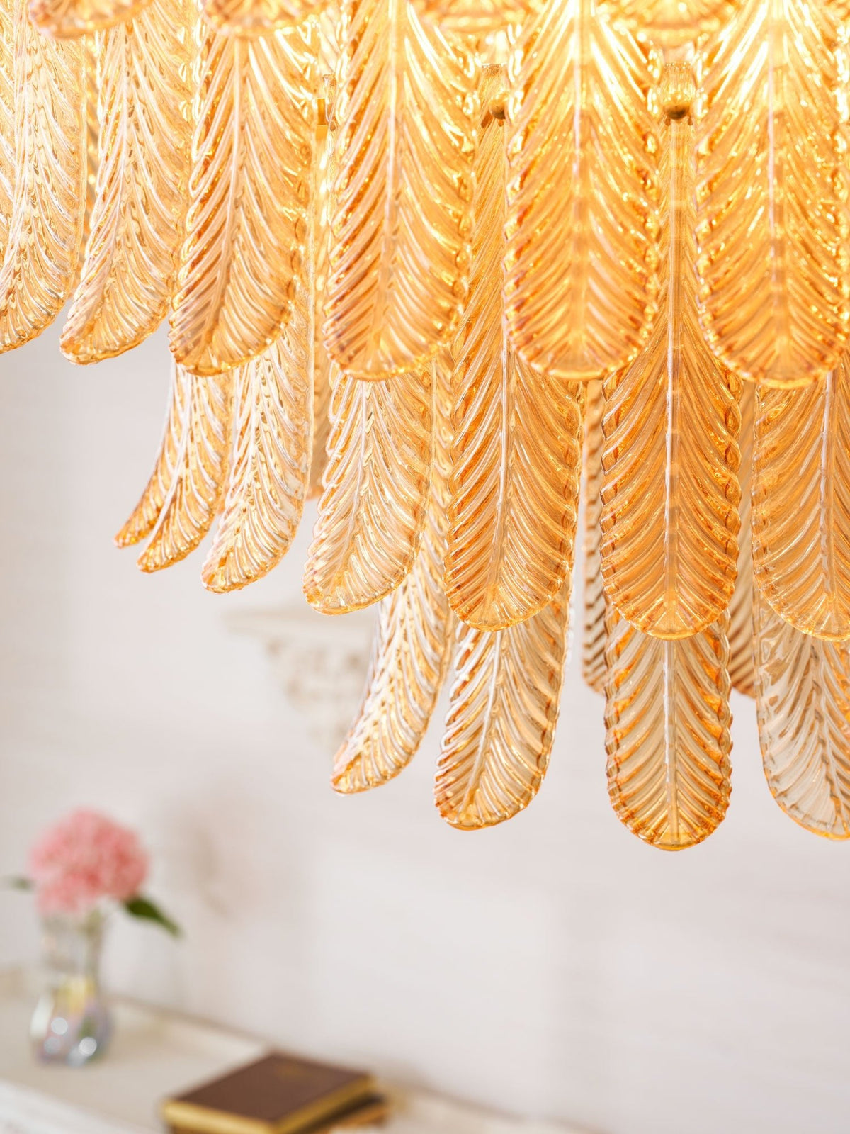 Close view of the overlapping amber glass leaves on this Clérachic pendant, with a vase of pink flowers blurred below.