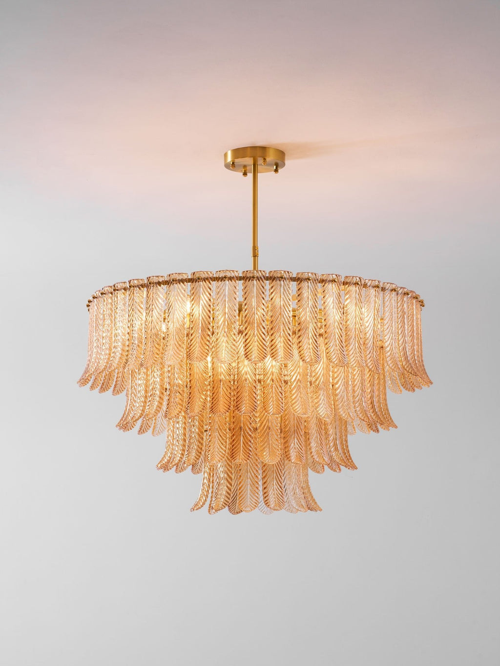 A Clérachic amber glass leaf pendant, lit, hangs from a brass rod and ceiling plate against a soft grey-pink backdrop.