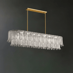 A Clérachic linear pendant of clear glass leaves hangs from a brass bar on two rods, against a dark green-grey wall.