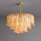 A lit Clérachic pendant of amber glass leaves in three tiers glows from the inside, hung from a brass rod on a grey backdrop.