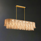 A Clérachic linear pendant of amber glass leaves hangs from a brass bar on two rods, switched off against a dark wall.
