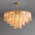 A wide, lit Clérachic pendant of amber glass leaves in tiers hangs from a brass rod and round ceiling plate on grey.