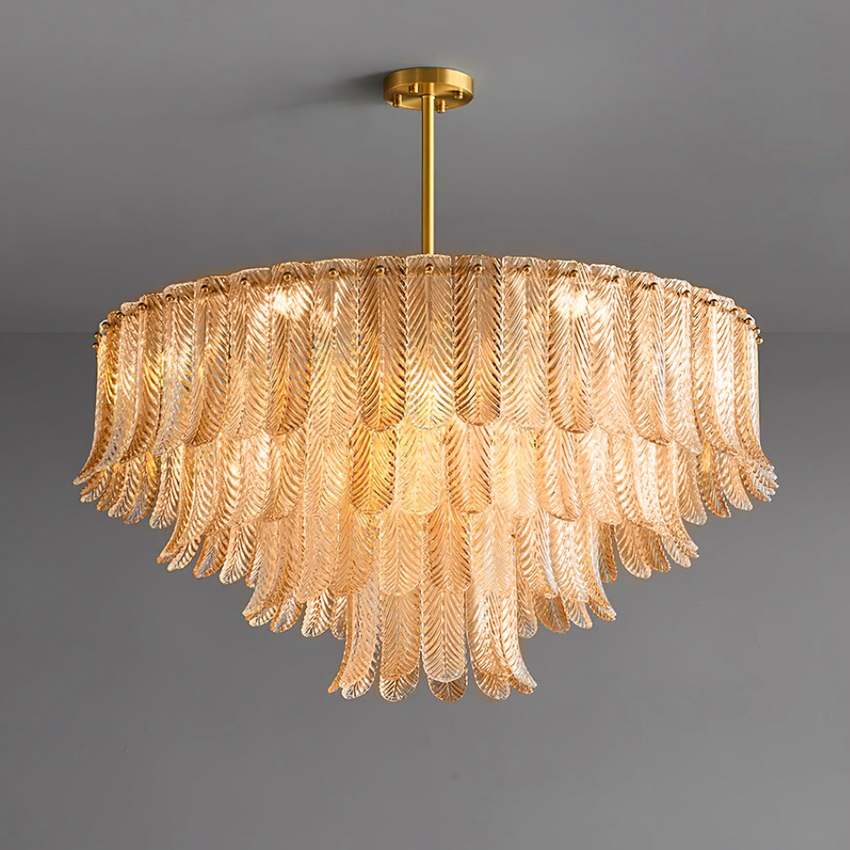 A wide, lit Clérachic pendant of amber glass leaves in tiers hangs from a brass rod and round ceiling plate on grey.