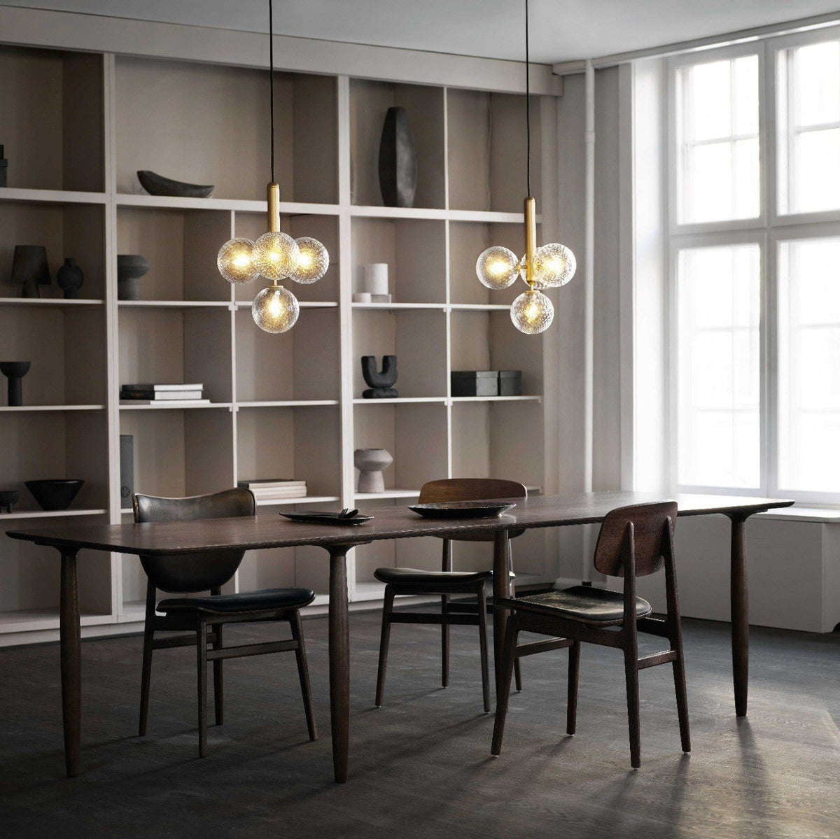Two Clérachic cluster pendants with clear glass globes hang beside tall white shelving over a dark wood dining table.