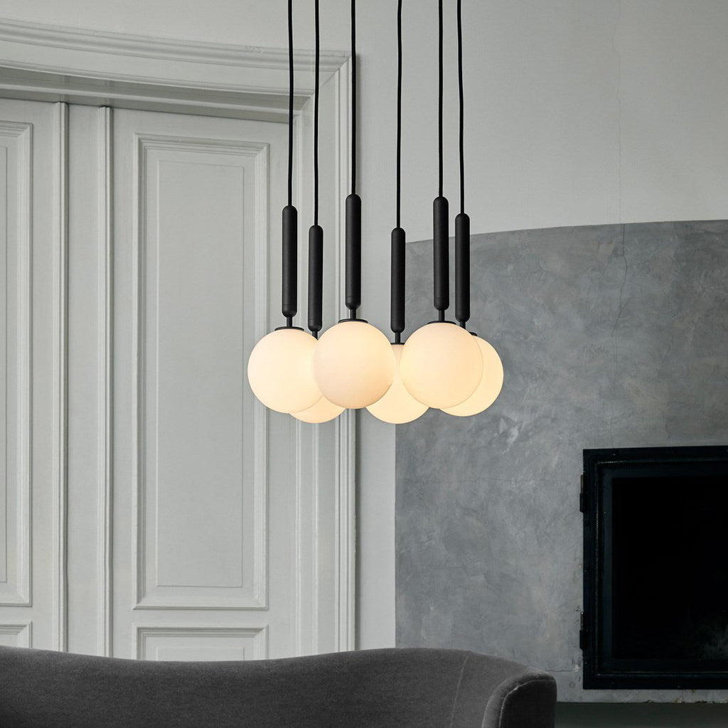 A group of black-rod Clérachic pendants with white opal globes hangs before a panelled wall and a grey plaster fireplace.
