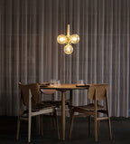 A brass cluster pendant from Clérachic glows amber above a round walnut table, with dark curtains behind.