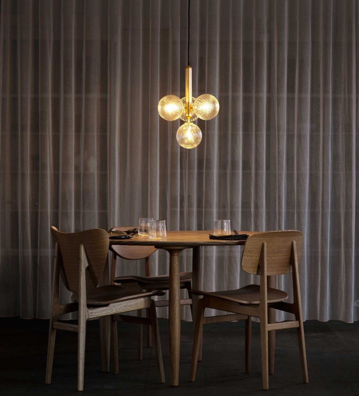 A brass cluster pendant from Clérachic glows amber above a round walnut table, with dark curtains behind.
