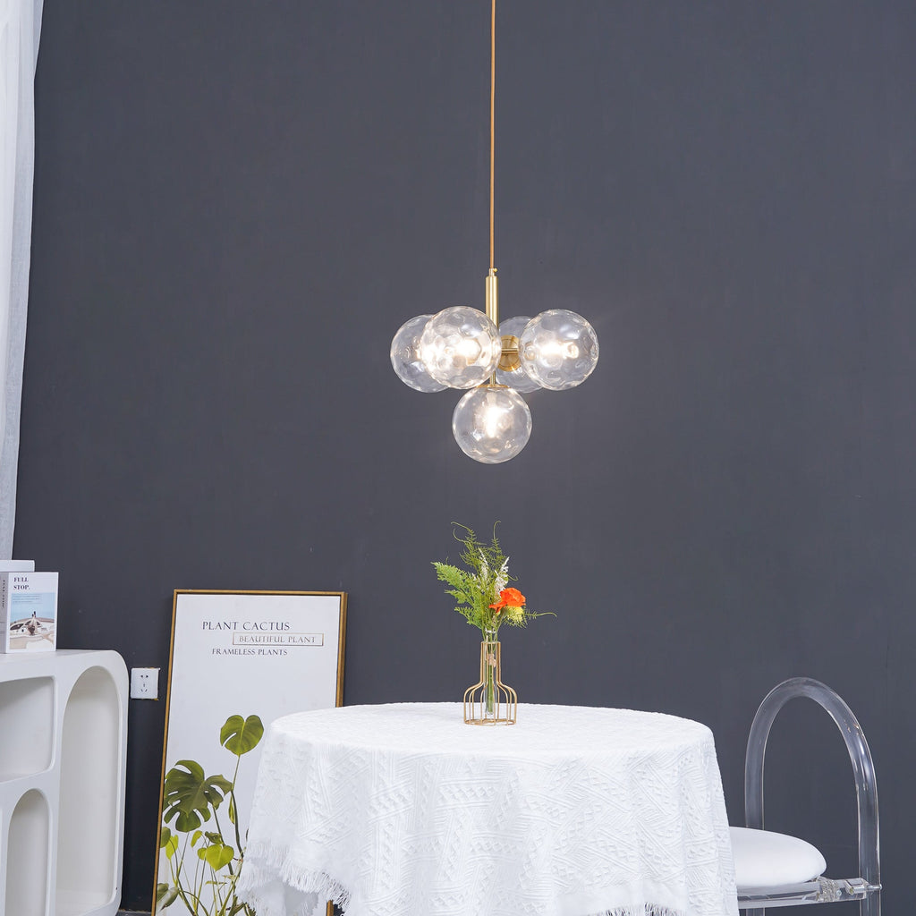 A gold-rod Clérachic cluster pendant with clear glass globes hangs against a dark grey wall above a white-clothed table.