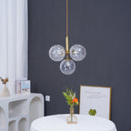 Gold-rod Clérachic pendant with three clear rippled glass globes against a dark grey wall, with a flower vase below.
