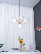 Five-globe Clérachic pendant with clear glass on a gold rod, hanging in front of white curtains and a round table.