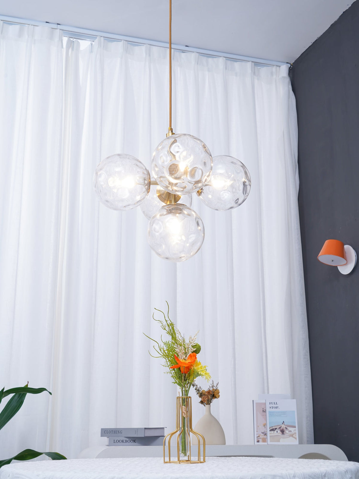 Five-globe Clérachic pendant with clear glass on a gold rod, hanging in front of white curtains and a round table.