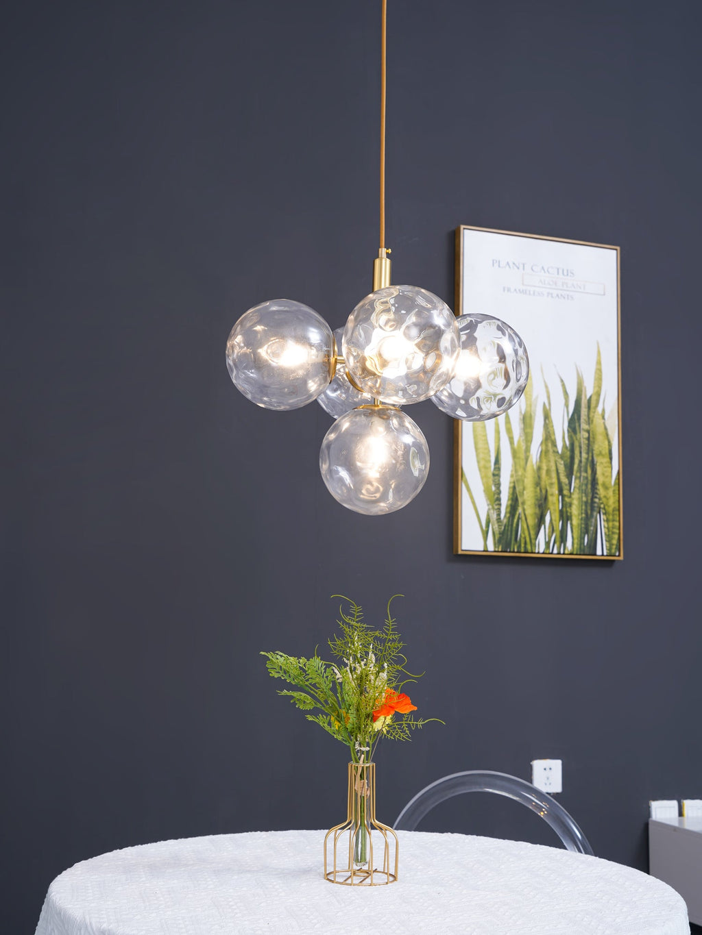 Gold-rod Clérachic cluster pendant with clear rippled globes and a lit bulb, hanging against a dark grey wall.