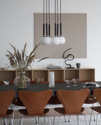 Five black-rod Clérachic pendants with opal globes hang together over a dark table and tan leather chairs.