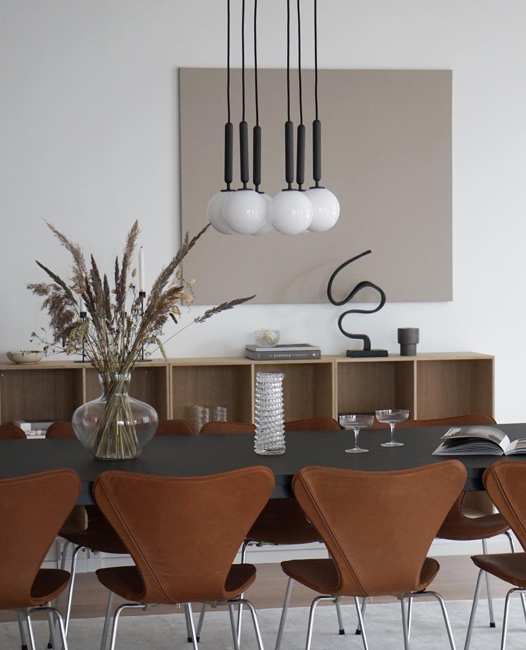 Five black-rod Clérachic pendants with opal globes hang together over a dark table and tan leather chairs.
