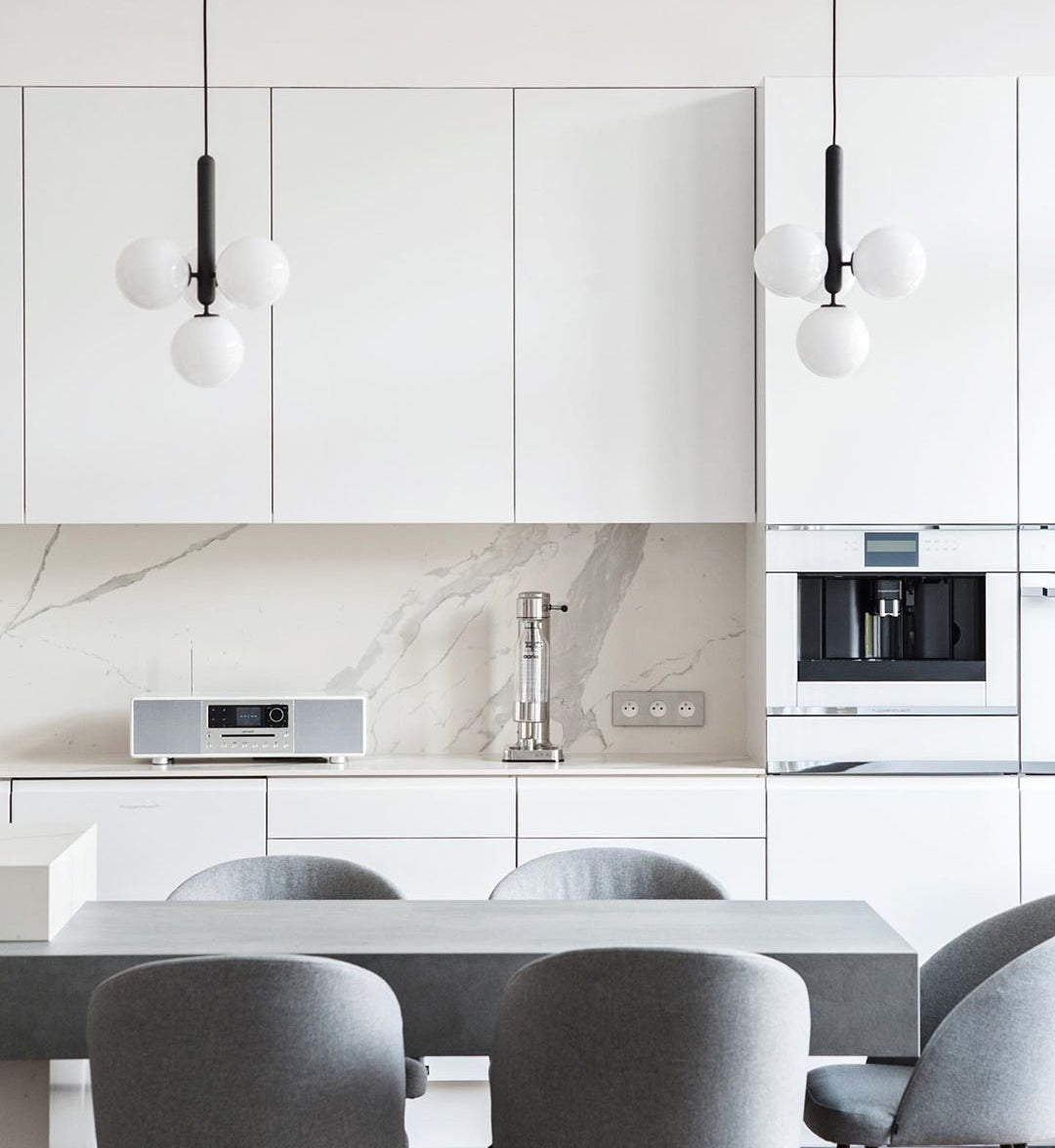Two white opal cluster pendants on black rods from Clérachic hang in a white kitchen with a marble splashback.
