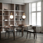 Two brass-rod Clérachic pendants with single white globes hang in front of open shelving and a dark dining table.