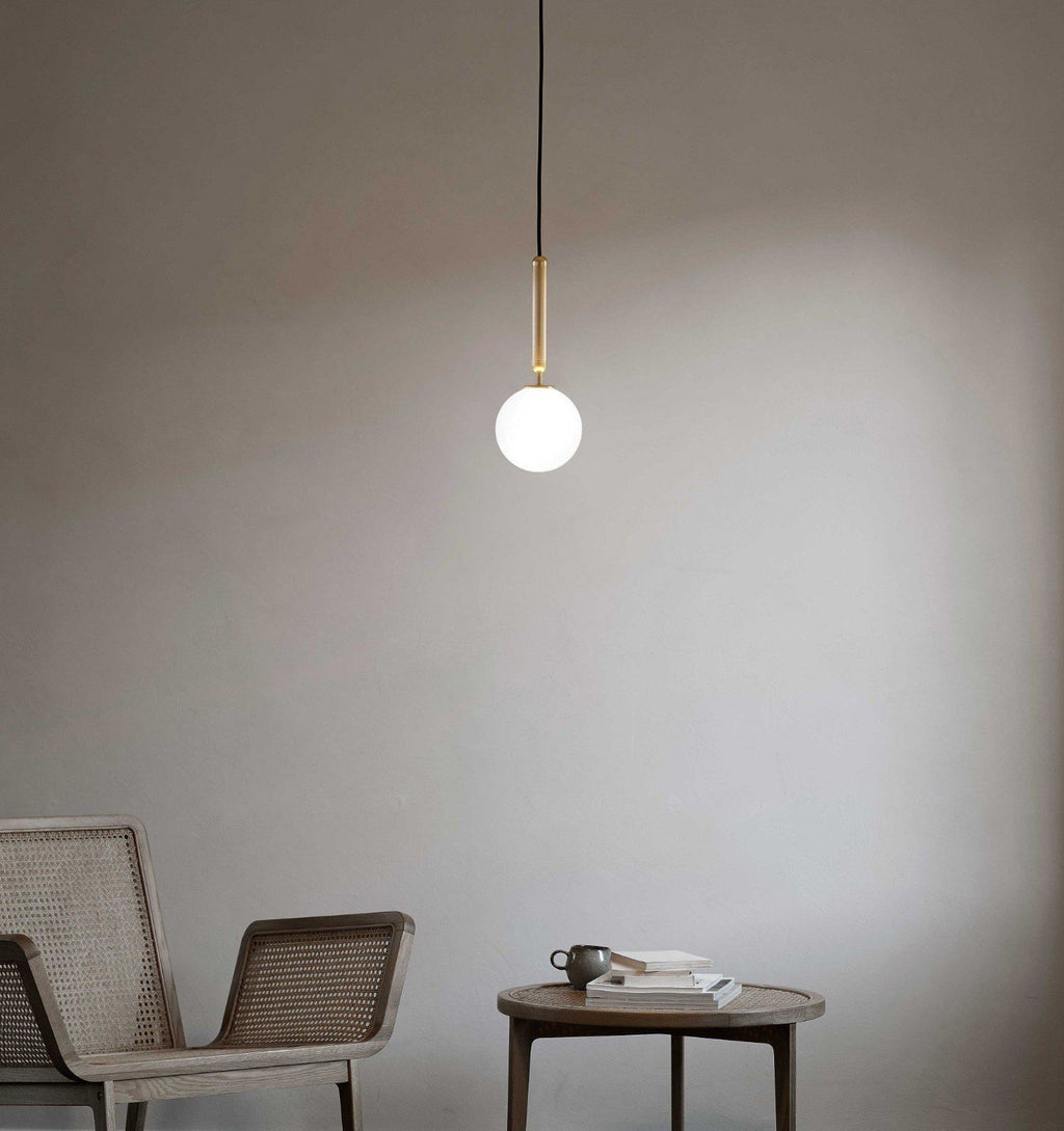A single brass-rod Clérachic pendant with a white opal globe hangs against a soft grey wall beside a cane chair.