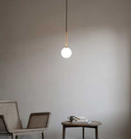 A single brass-rod Clérachic pendant with a white opal globe hangs against a soft grey wall beside a cane chair.