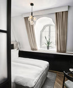 A black-rod Clérachic pendant with clear glass globes hangs in a hotel-style bedroom with beige curtains.