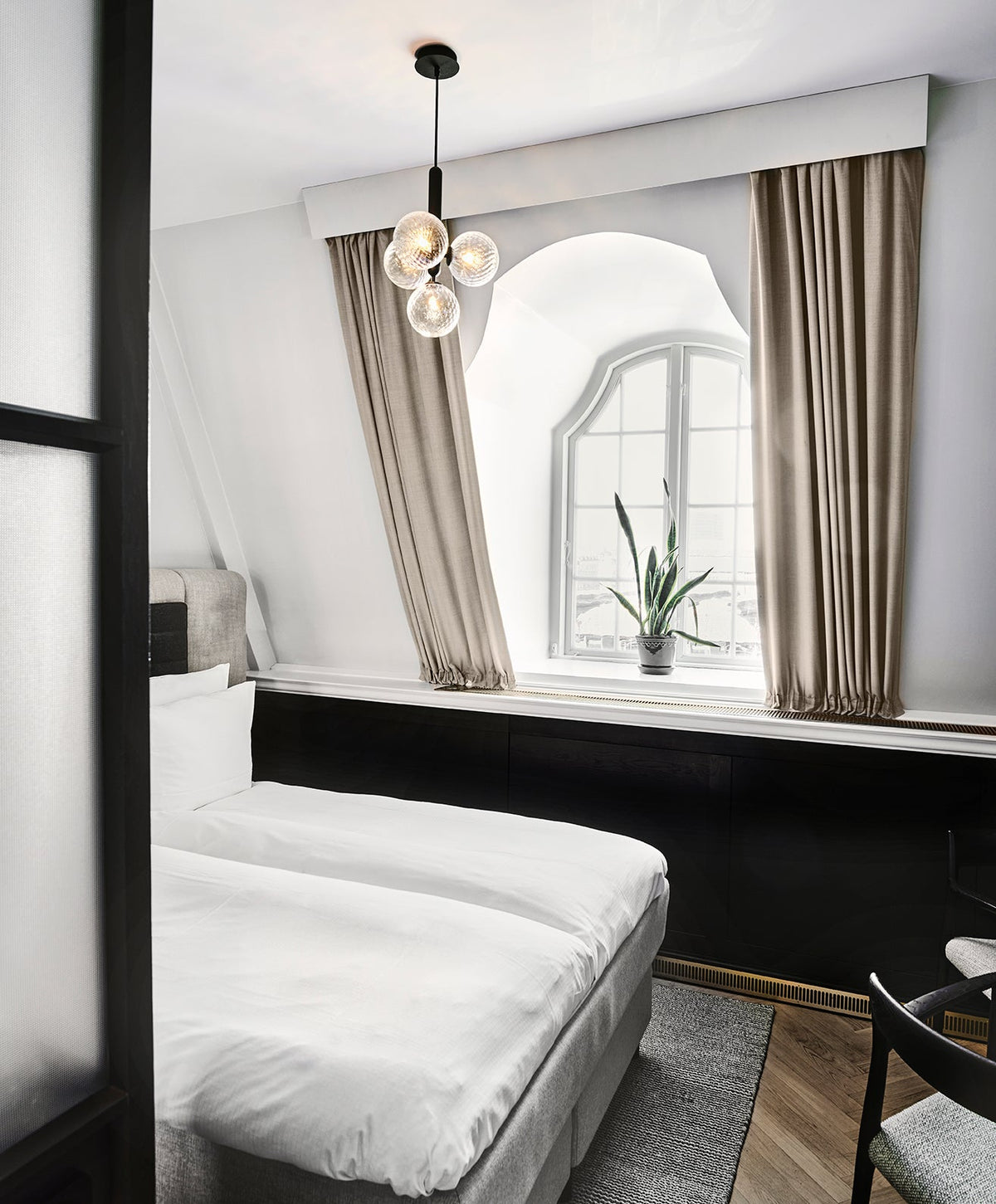 A black-rod Clérachic pendant with clear glass globes hangs in a hotel-style bedroom with beige curtains.