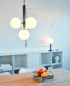 Black-rod Clérachic pendant with three large opal globes lit warm, hanging above a table in a pale blue room.