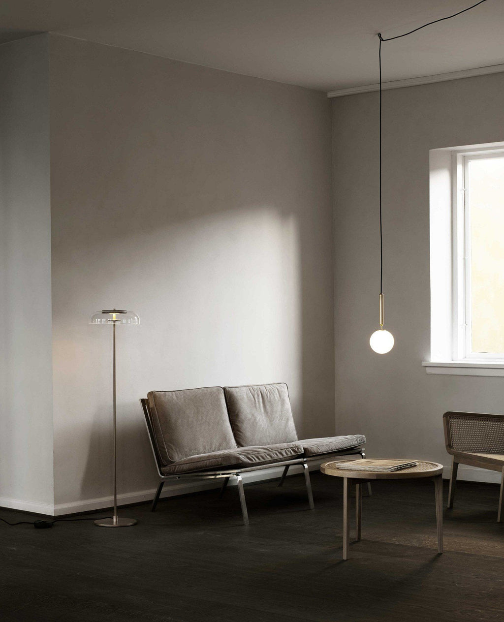 A single-globe Clérachic pendant on a long cord hangs low beside a sofa and floor lamp in a quiet grey room.