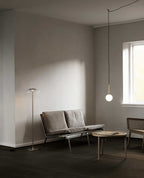 A single-globe Clérachic pendant on a long cord hangs low beside a sofa and floor lamp in a quiet grey room.