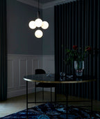 Four opal globes glow on a black-rod Clérachic pendant above a dark round table, with navy curtains behind.