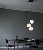 Three black-rod Clérachic pendants with opal globes hang at different heights above a dark table in a grey room.