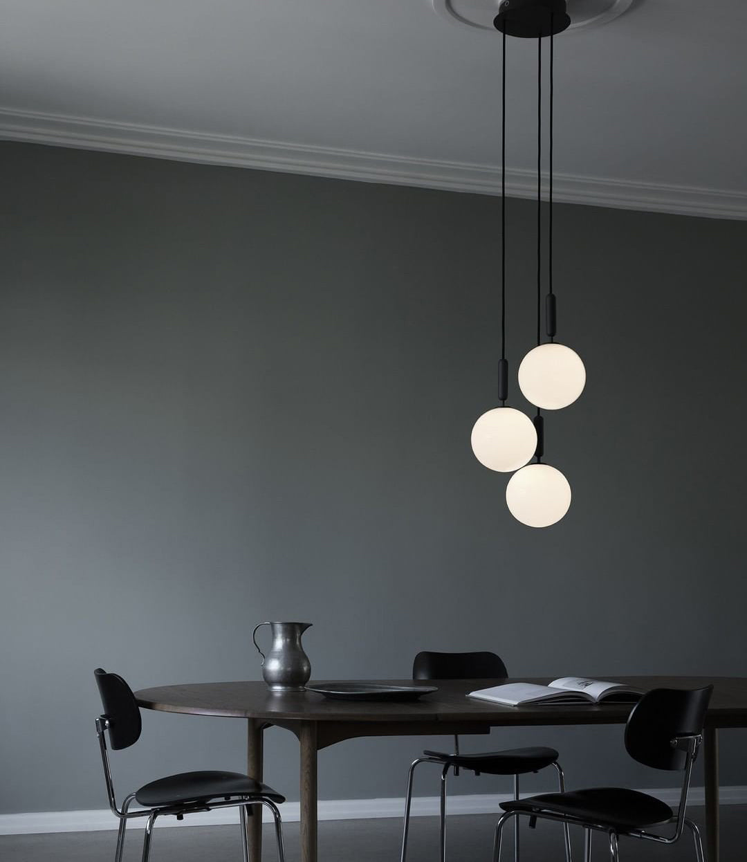 Three black-rod Clérachic pendants with opal globes hang at different heights above a dark table in a grey room.