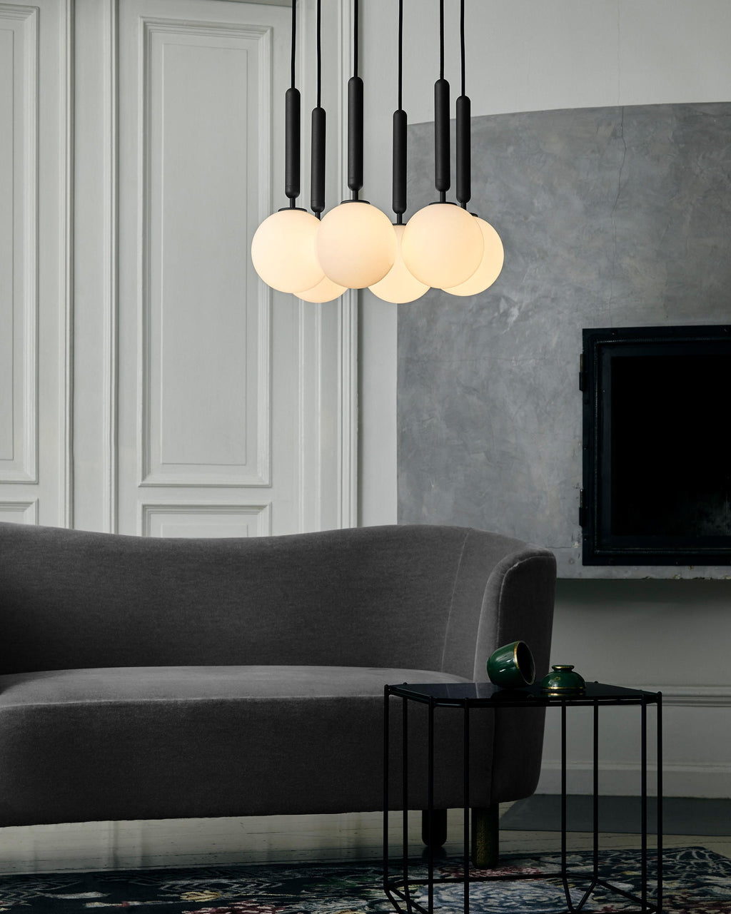 Black-rod Clérachic pendants with warm-lit opal globes hang above a grey sofa in front of a panelled wall.