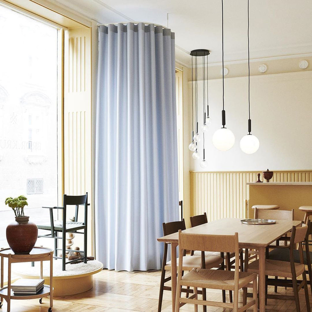 Several Clérachic pendants in brass and white opal glass hang over a dining table in a room with pale blue curtains.
