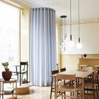 Several Clérachic pendants in brass and white opal glass hang over a dining table in a room with pale blue curtains.
